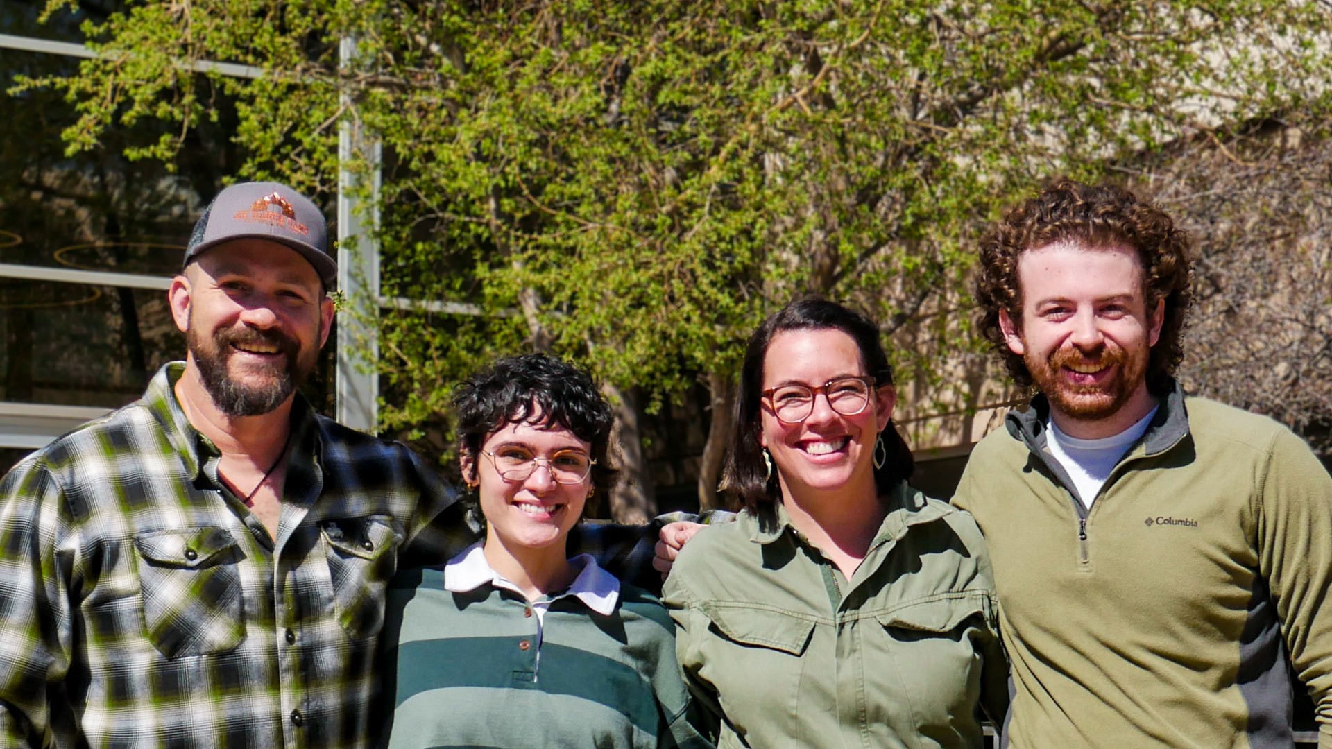 four people standing outside the JCC, smiling. Two men on the outside and two women. Most wear green to represent their membership on the Green Team