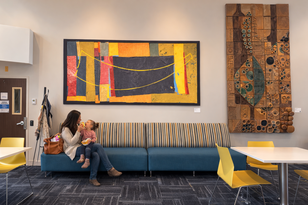 A woman and child sit in the lobby of the JCC on a bench below the Shofar artwork.